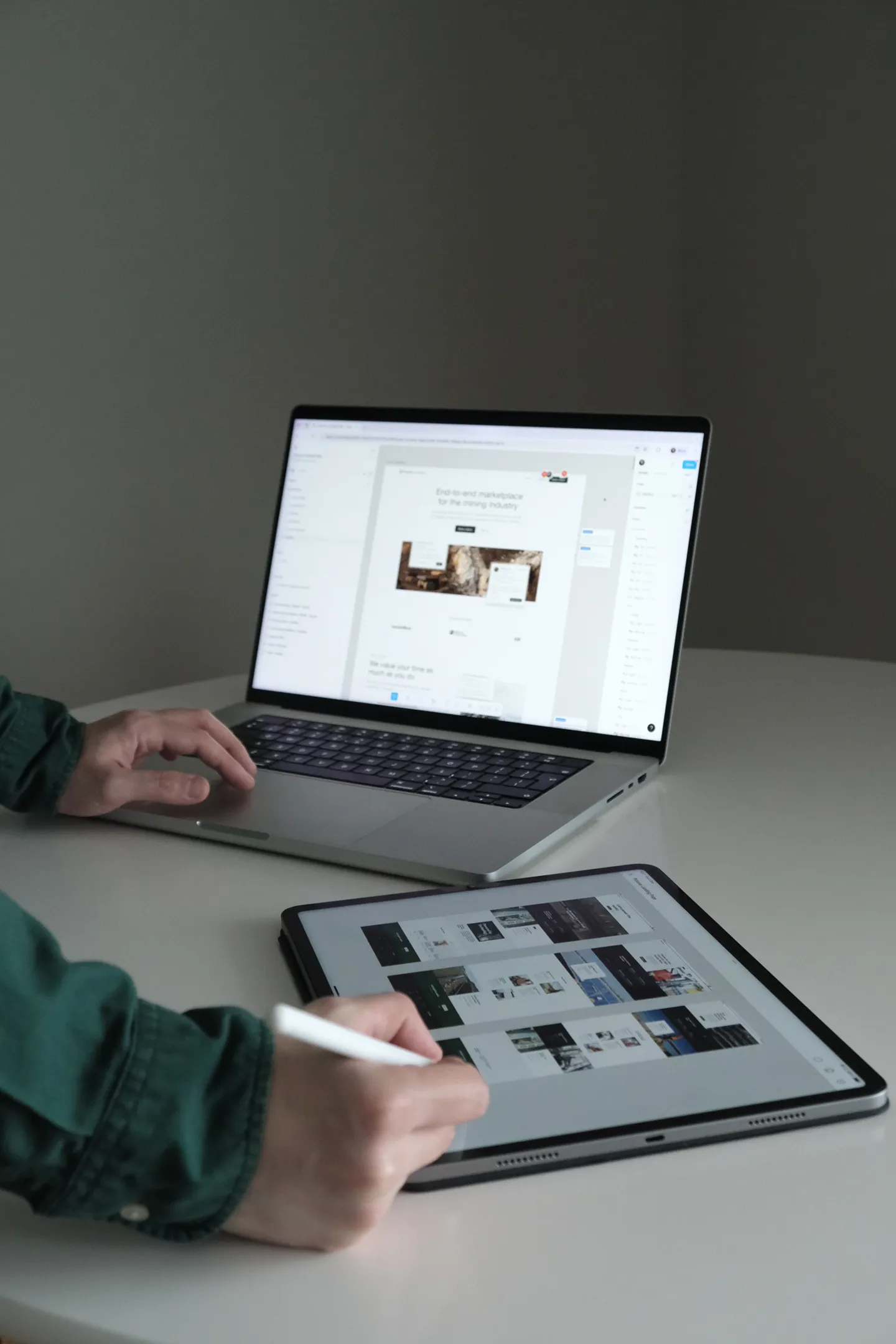 Persoană care folosește un stylus pe o tabletă cu machete de design, iar în fundal un laptop afișează designul unei pagini web.