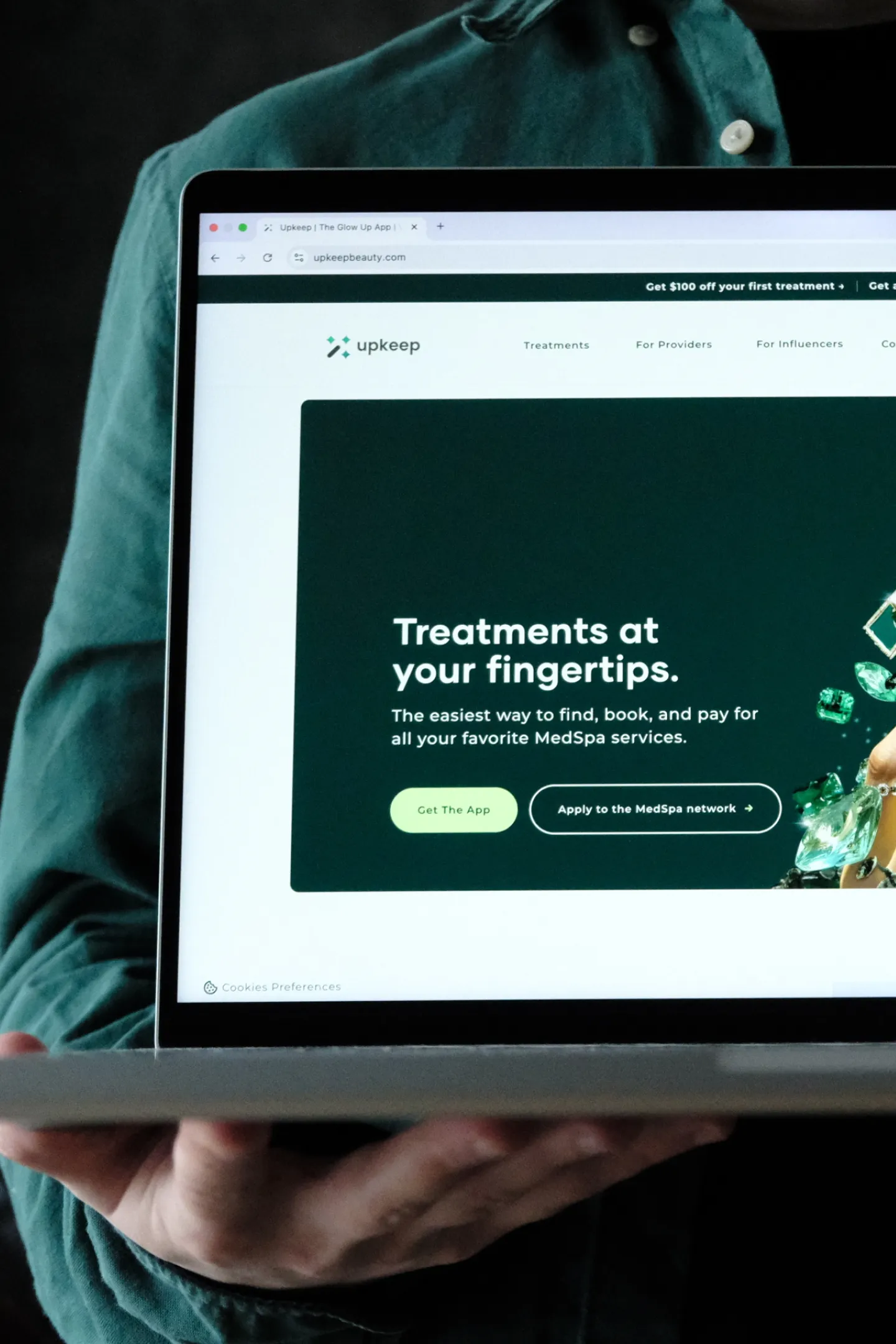 Persoană în cămașă verde ținând un laptop pe care se vede aplicația Upkeep pentru rezervarea tratamentelor MedSpa.