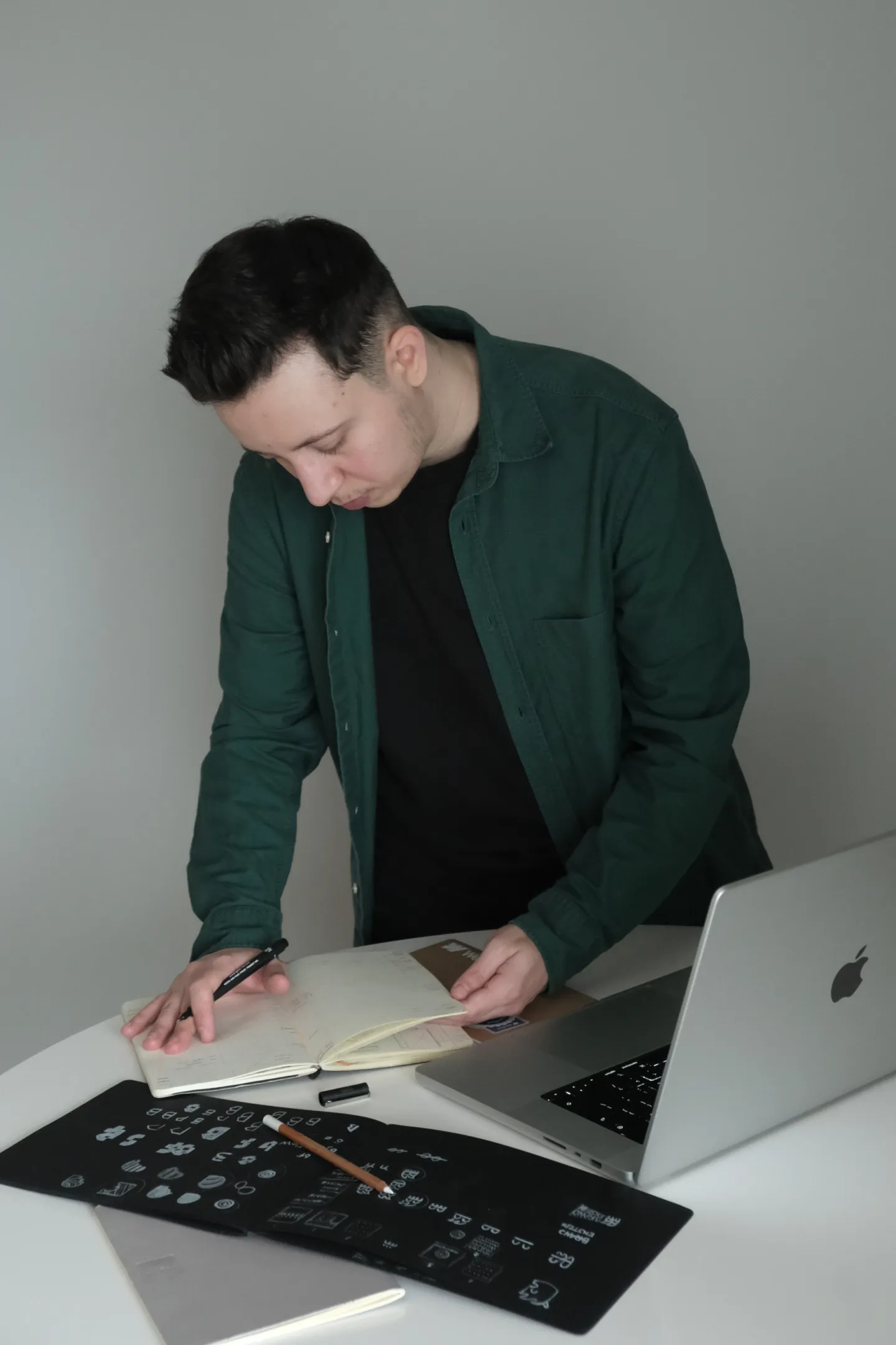Tânăr în cămașă verde care scrie într-un caiet la un birou, cu laptop și schițe de design.